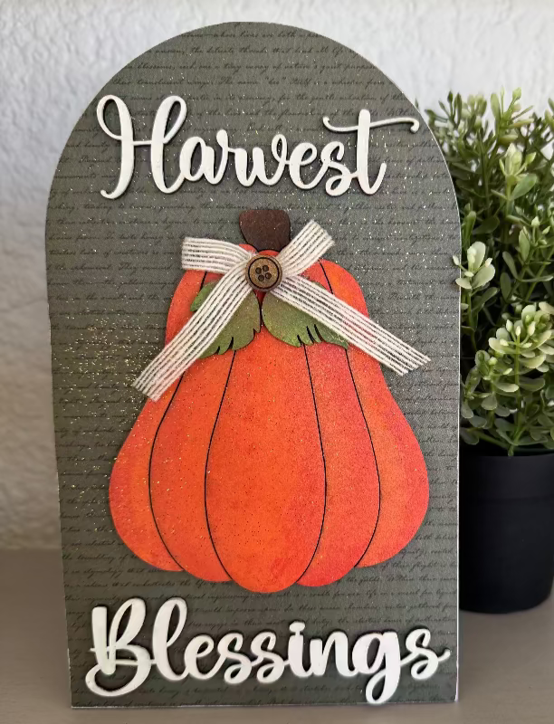 Decorative sign with a pumpkin and 'Harvest Blessings' text, placed on a surface with a plant in the background.